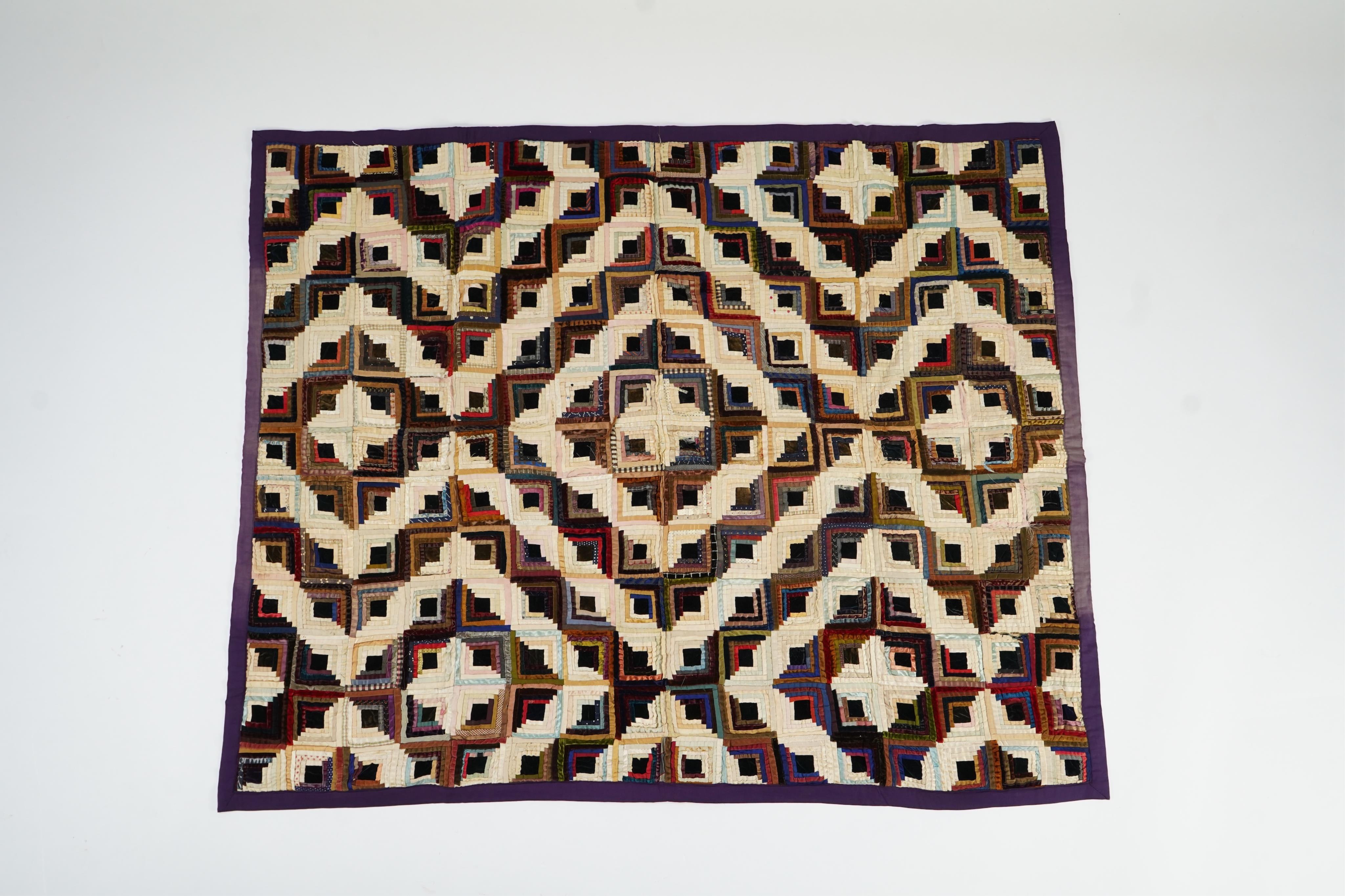 An early 20th century silk and velvet ‘Log Cabin’ designed bed cover, with a deep purple cotton backing, 140cm x 74cm. Condition - various thin strips have torn or are worn from constant use, so needs restoration.